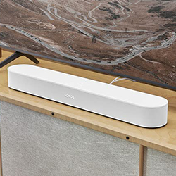 White soundbar on a wooden TV stand, beneath a wall-mounted television showing a landscape image. The scene conveys a modern and sleek home entertainment setup.