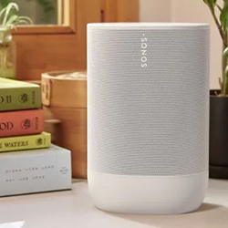 A sleek, grey Sonos speaker on a wooden surface near a stack of colourful books and a window. The scene is calm and tidy, suggesting a cosy, modern home.