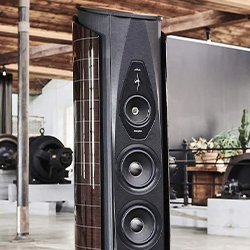 A sleek, modern black speaker with three prominent circular drivers in a stylish, rustic room. The ambiance is warm and sophisticated.