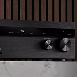 A sleek black audio receiver with two large knobs and a digital display reading "TV." It's set against a wood panel background, conveying a modern, elegant feel.