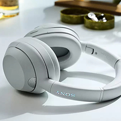 Sleek white over-ear headphones rest on a sunlit table. A wooden tray and small plant in the background add a modern, calm ambiance.