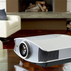 A sleek, modern projector rests on a glossy coffee table in a cozy living room. A white sofa and fireplace create a warm, inviting atmosphere.