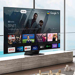 A modern living room with a large flat-screen TV displaying the Google TV interface. Apps like Netflix and YouTube are visible. The room has floor-to-ceiling windows overlooking a serene ocean view.