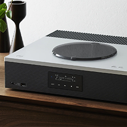 A sleek, modern audio system on a wooden surface. A vinyl record player is prominent, with a minimalistic design alongside a small plant and vase.