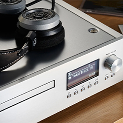 Silver audio amplifier with a digital display showing "Total Track: 19" and large headphones resting on top. The scene evokes a modern, sleek vibe.