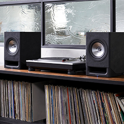 A vinyl record player flanked by two black speakers sits on a wooden shelf. Below, a collection of vinyl records is neatly arranged, evoking a retro vibe.