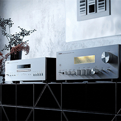 Two vintage silver stereo receivers on a black tiled shelf, with one displaying a lit panel. A small potted plant adds a decorative touch, creating a retro vibe.