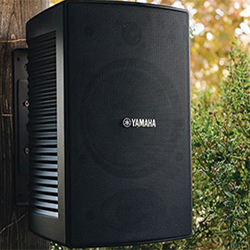 Outdoor black Yamaha speaker mounted on a wooden post, surrounded by greenery and trees, conveying a serene and natural auditory setting.