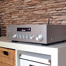A silver stereo receiver with dials and display sits on a white shelf, against a textured stone wall, conveying a modern and sleek atmosphere.