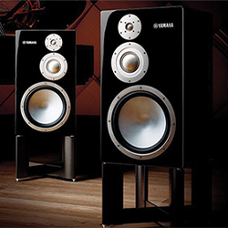 Two sleek Yamaha speakers on a wooden floor. Each black speaker has three silver circular components, reflecting a modern, high-quality audio setup.
