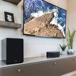 Modern living room with a wall-mounted TV displaying a scenic ocean view. Below, a sleek cabinet holds a speaker, décor items, and a small plant.