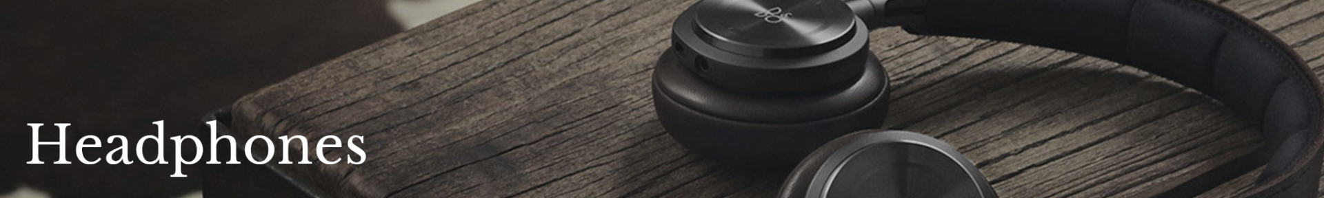 Close-up of black over-ear headphones resting on a rustic wooden surface. The word "Headphones" is written in white on the left side. The tone is modern and stylish.