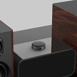 A modern audio setup with a central stereo system and two speakers on a white cabinet. A person holds a red smartphone in the foreground.