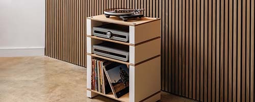 A sleek stereo system with speakers and receiver on a minimalist shelf, beside a modern lamp and soft fur throw, set against a concrete wall.