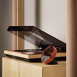 A sleek, modern turntable with a transparent dust cover sits on a wooden cabinet. A small, round, amber-hued controller complements the stylish setup.