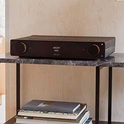 A sleek black audio receiver sits on a marble shelf, surrounded by stacked books. The display reads "INPUT A25." The setting is minimal and modern.