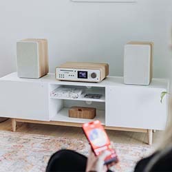A modern audio setup with a central stereo system and two speakers on a white cabinet. A person holds a red smartphone in the foreground.