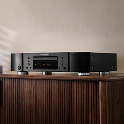 A sleek black audio component with a CD tray sits on a wooden cabinet against a neutral wall, conveying a minimalist and modern vibe.
