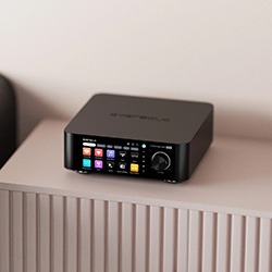 A sleek black electronic device with a colourful digital display and a control knob sits on a textured light pink surface.
