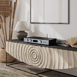 Simple, modern living room with a textured wooden sideboard holding a black audio system, ceramic lamp, and open book.