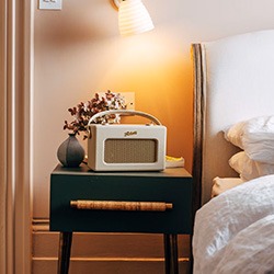 A cosy bedroom scene with a retro radio on a dark bedside table, soft lamp light, a small vase with flowers, and a white bed.