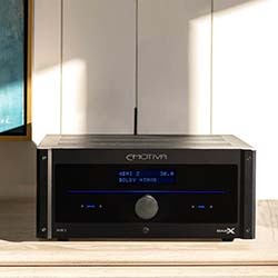 Black audio receiver on a wooden surface, displaying blue LED text "Dolby Atmos." Sunlight casts a warm glow on the scene.