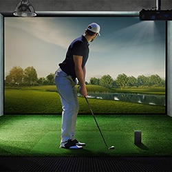 A man is poised to hit a golf ball in an indoor simulator, featuring a realistic golf course projected on a screen. The scene is calm and focused.