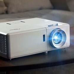 A white projector emits a bright blue light from its lens, sitting on a wooden surface. A remote control lies nearby, suggesting a setup for a presentation.