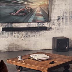 A cosy living room features a TV with a racing car scene, wooden coffee table with books and a cup, soundbar, and speaker, conveying a modern, relaxed vibe.