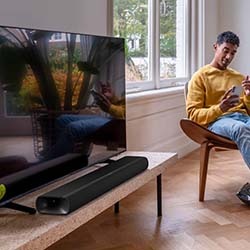 A man in a cosy living room setting sits on a stylish wooden chair, smiling at his phone. A sleek TV and soundbar are on a wooden table, with large windows framing a leafy garden view.