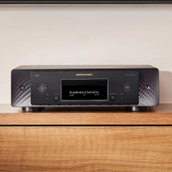 A sleek black audio receiver with a digital display showing "Sound Group Network" sits on a wooden shelf against a neutral wall, conveying a modern, minimalistic vibe.