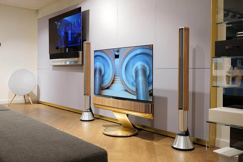 Modern living room with two futuristic TVs mounted on a wall, wooden floors, and sleek speakers. The ambiance is sleek and high-tech.