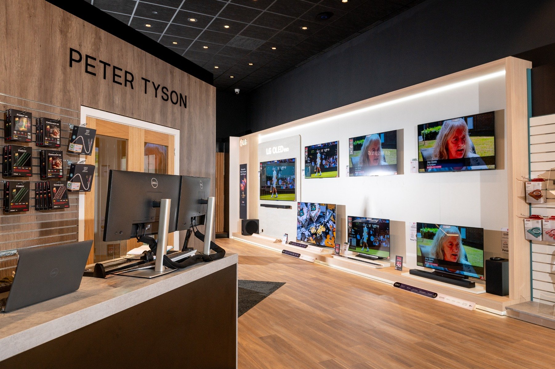 Electronics store interior with wooden floors, featuring multiple LG OLED TVs playing sports and news. A counter has two Dell monitors and a laptop.