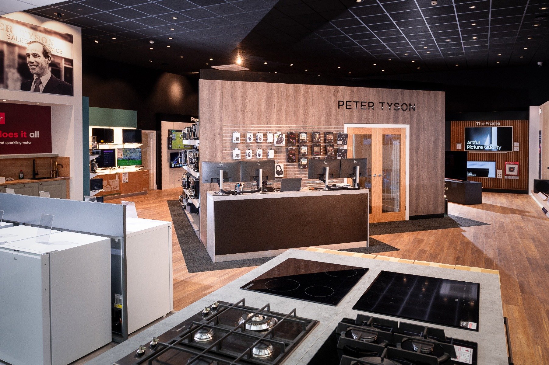 Modern electronics store interior with wood accents and digital displays. Features kitchen appliances, a sales counter with computers, and tech products.