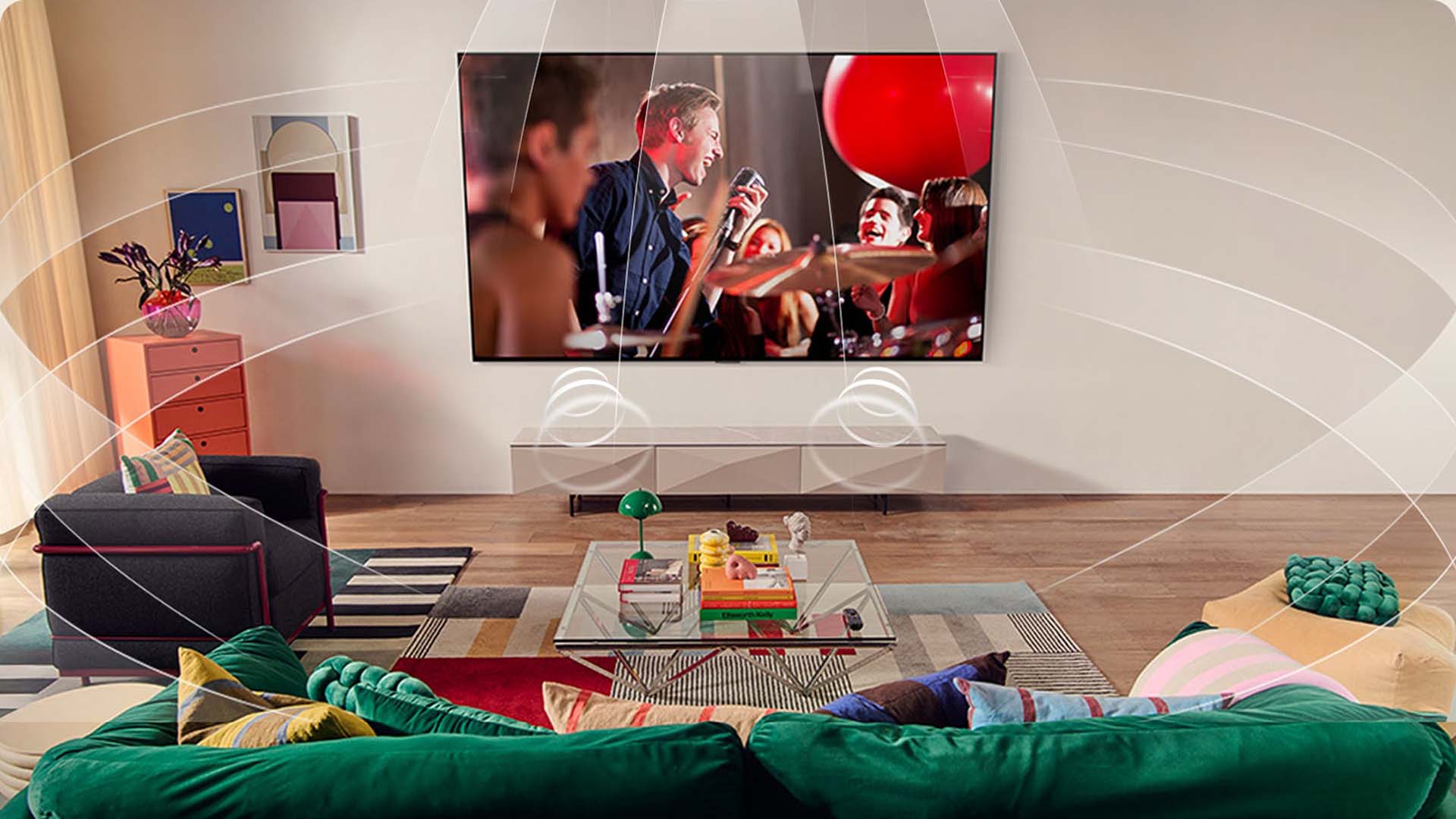 A cozy living room with vibrant décor features a large wall-mounted TV showing a lively concert. Green velvet sofa and colourful cushions add warmth.