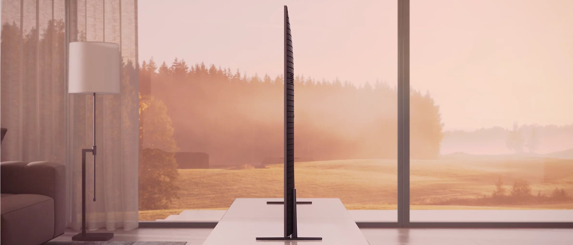 Side view of TV on a table in a living room showing slim design with view through a window of sunrise over a garden in background.