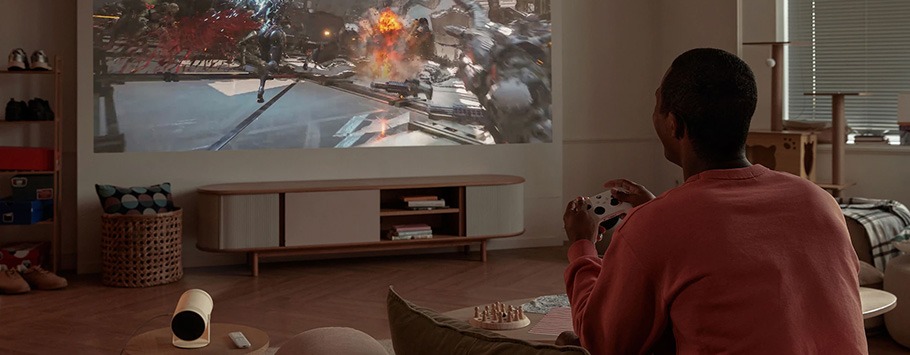 Person playing a video game on a large projection screen in a cosy living room. The room features warm lighting, wooden furniture, and a chessboard.