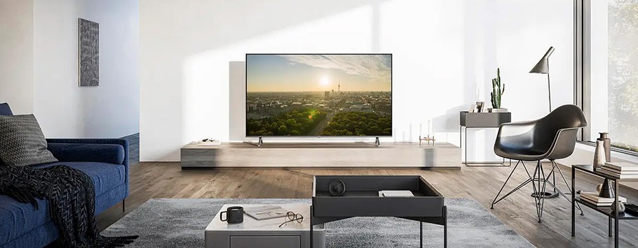 Modern living room with a sleek TV displaying a sunset over a cityscape. Blue sofa, black chair, and minimalist décor create a serene ambiance.