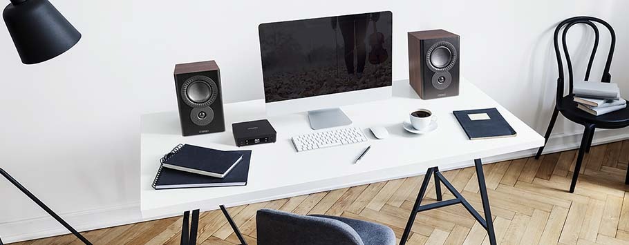 A minimalist workspace features a large monitor, two speakers, keyboard, and mouse on a white desk. A chair, books, and a cup add a cosy touch.