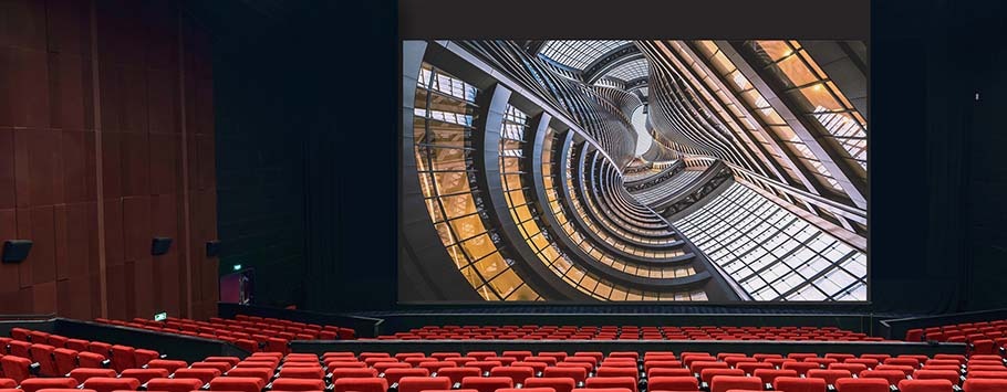 A modern theatre with red seats faces a large screen displaying a spiral architectural design. The atmosphere is dramatic and futuristic.