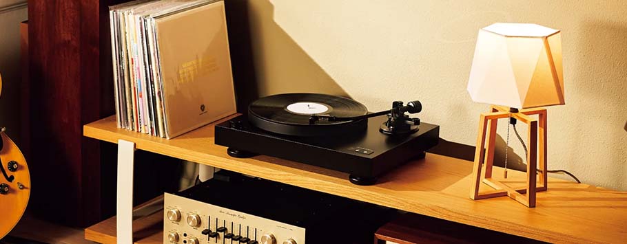 Warmly lit room with a wooden shelf holding a record player, vinyl records, a retro amplifier, and a modern geometric lamp.