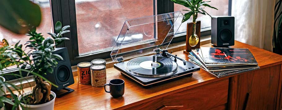 A cozy scene with a wooden console holding a vinyl record player, spinning a record, flanked by small plants, speakers, and a stack of albums near a window.