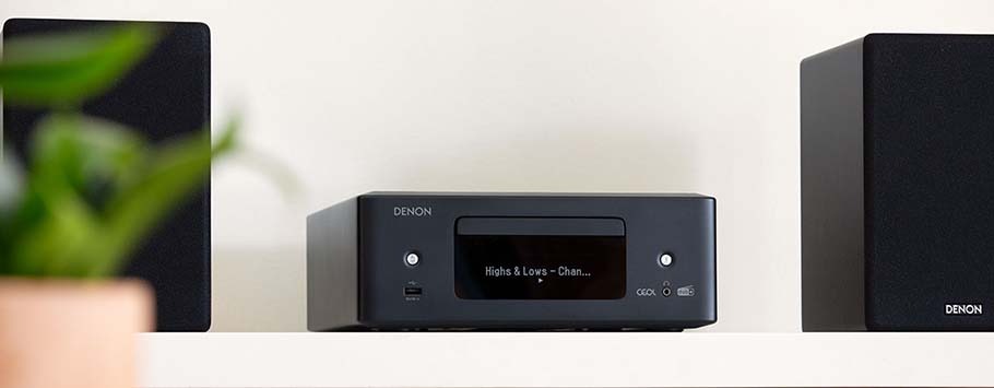 Black audio system with central display reading "Highs & Lows" between two speakers on a white shelf. A blurred plant is in the foreground.