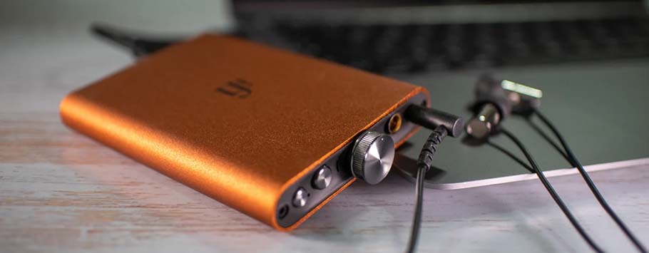 An orange portable audio device with a volume knob and ports sits on a wooden surface, connected by cables to a laptop.