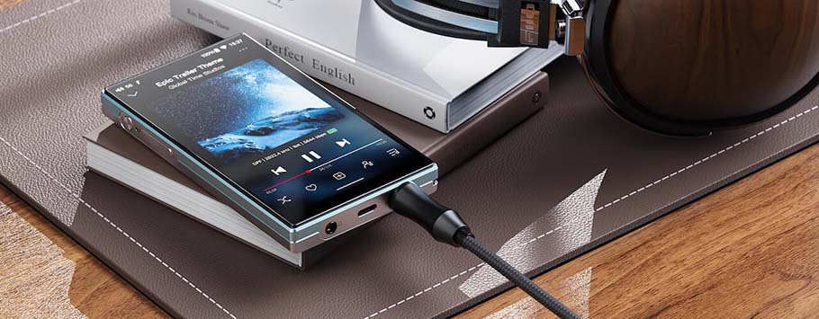 A portable music player displaying a song is placed on a closed book, next to over-ear headphones on a leather surface.
