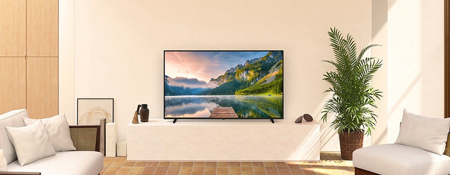 Modern living room with a wall-mounted TV showing a serene mountain lake scene. Minimalist furniture, green plant, neutral tones, and a cosy vibe.
