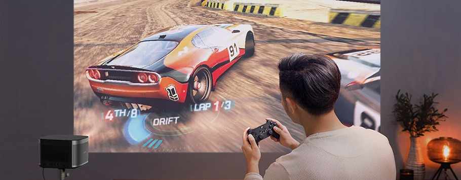 A person plays a racing video game projected on a wall. The game shows a car drifting on a track. The setup includes a small projector and cosy lighting.