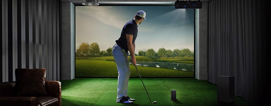 A man in a golf stance inside a virtual golf simulator room with a large screen displaying a scenic golf course, creating a focused and immersive atmosphere.