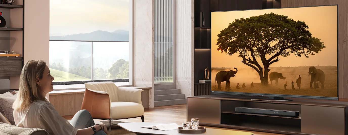 A woman sits on a sofa, smiling, watching a large TV displaying a serene savanna scene with elephants under a tree. The room is modern and bright.
