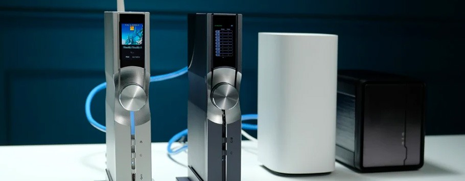 Two sleek, vertical audio amplifiers with round knobs and small screens stand on a white table. In the background are a white router and a black server.